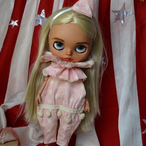 Op de afbeelding: Een blonde pop met een roze en witte sterrenprint jumpsuit met een witte ruchekraag en een roze en witte sterrenprint hoed. De pop staat voor een rood-wit gestreept doek met witte sterren.
