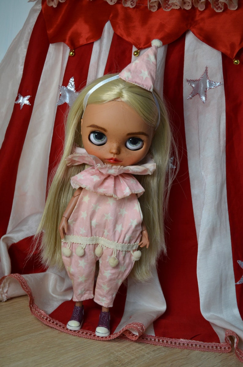 Op de afbeelding: Een blonde pop met een roze en witte sterrenprint jumpsuit met een witte ruchekraag en een roze en witte sterrenprint hoed. De pop staat voor een rood-wit gestreept doek met witte sterren.