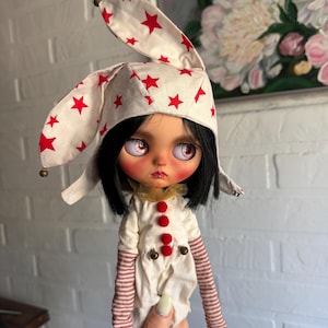Può includere: Una bambola con capelli neri e occhi viola chiaro che indossa un cappello bianco con stelle rosse e un completo bianco con pon pon rossi, piccoli campanelli metallici e maniche lunghe a righe orizzontali rosse e bianche.