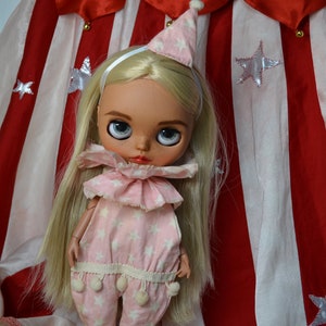 Op de afbeelding: Een blonde pop die een roze en witte sterrenprint jumpsuit draagt met een witte geruite kraag en een witte punthoed. De pop staat voor een rood en wit gestreepte stoffen achtergrond.
