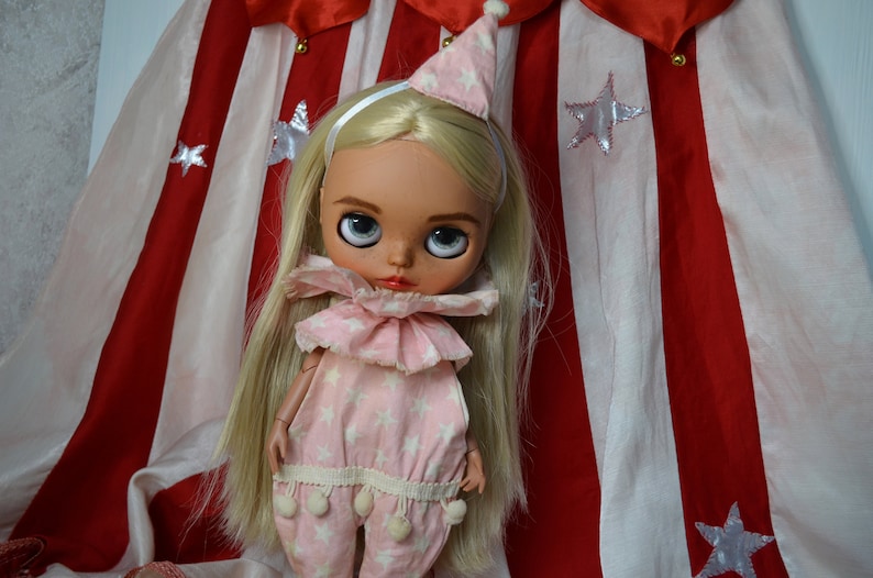 Op de afbeelding: Een blonde pop die een roze en witte sterrenprint jumpsuit draagt met een witte geruite kraag en een witte punthoed. De pop staat voor een rood en wit gestreepte stoffen achtergrond.