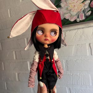 Pu&ograve; includere: Una bambola con capelli neri e grandi occhi chiari indossa un cappello da giullare rosso e bianco con piccoli campanelli. La bambola indossa un gilet nero e rosso con stelle dorate, una camicia bianca e maniche a righe, con pantaloni neri.