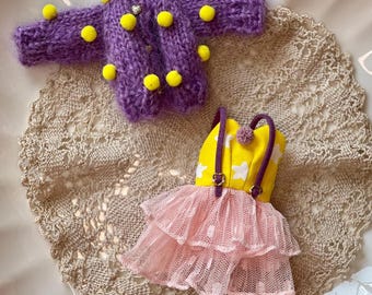 May include: A miniature purple knit sweater with yellow pom-poms and a yellow and pink dress with white star accents. The dress has a yellow top with purple straps and a pink tulle skirt. The items are displayed on a lace doily.