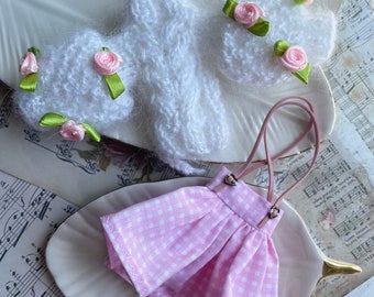 May include: A set of doll clothes featuring a pink and white gingham dress with straps and a white knitted hat with pink flowers.