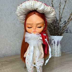 May include: A red-haired doll with closed eyes wearing a white dress with red polka dots and a white mushroom-shaped hat with a red trim.