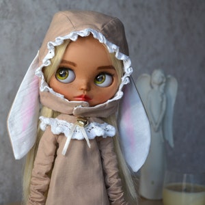 Puede incluir: Una mu&ntilde;eca con cabello rubio que lleva un vestido beige con capucha, ribete de encaje blanco y orejas de conejo. La mu&ntilde;eca tiene grandes ojos verdes y una cara pintada.