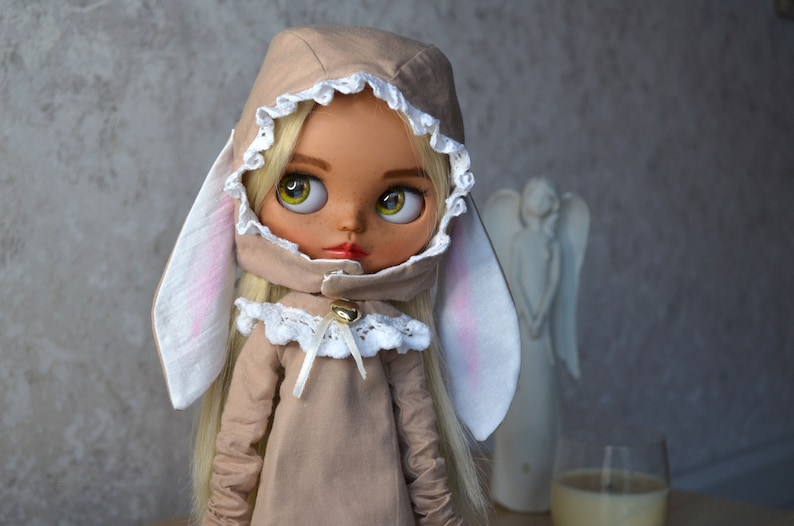 Puede incluir: Una mu&ntilde;eca con cabello rubio que lleva un vestido beige con capucha, ribete de encaje blanco y orejas de conejo. La mu&ntilde;eca tiene grandes ojos verdes y una cara pintada.