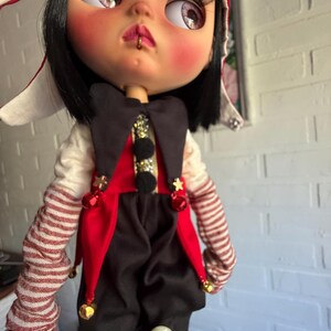 Pu&ograve; includere: Una bambola con capelli neri, ciglia lunghe e un piccolo anello dorato al labbro. Indossa un costume da giullare rosso e nero con stelle dorate, campanellini e maniche a righe. Un cappello rosso e bianco copre la testa e ha guance rosate.