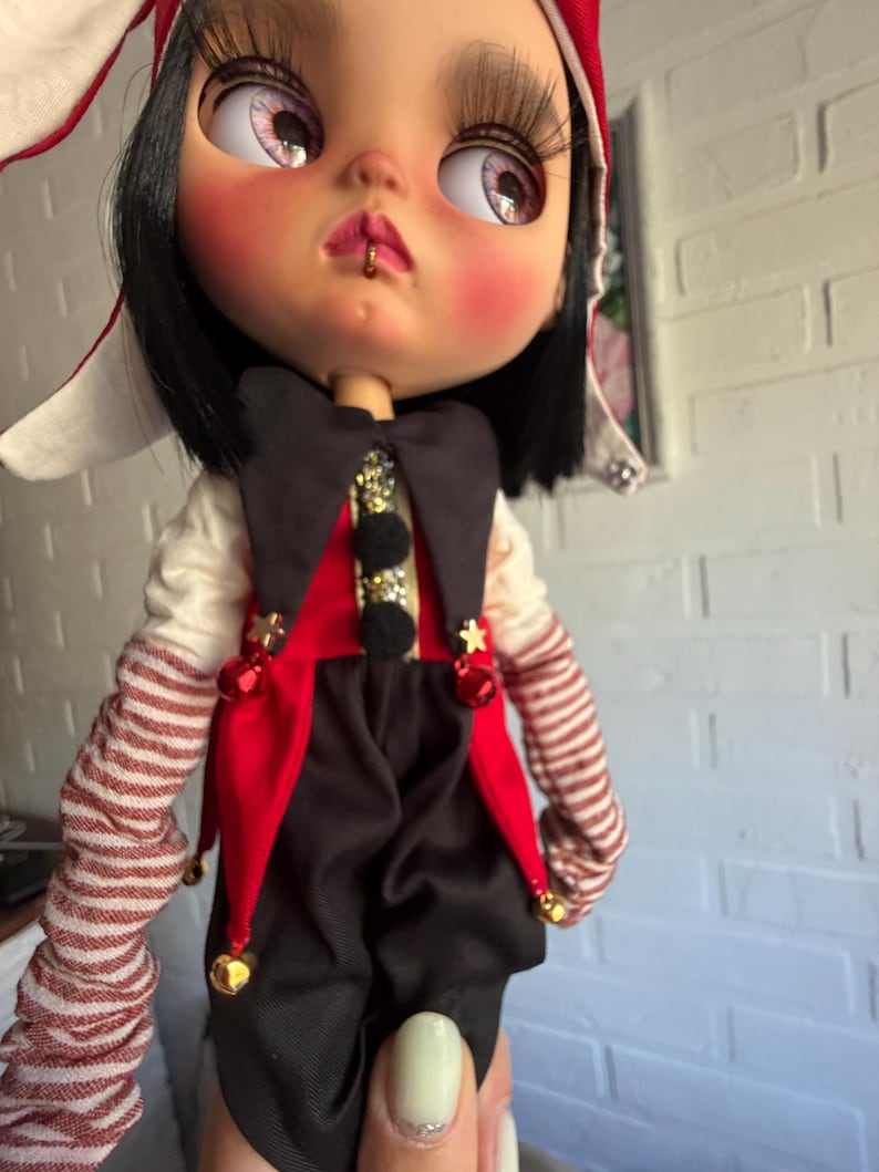 Pu&ograve; includere: Una bambola con capelli neri, ciglia lunghe e un piccolo anello dorato al labbro. Indossa un costume da giullare rosso e nero con stelle dorate, campanellini e maniche a righe. Un cappello rosso e bianco copre la testa e ha guance rosate.