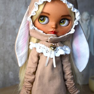 Puede incluir: Una mu&ntilde;eca con cabello rubio que lleva un vestido beige con capucha, ribete de encaje blanco y orejas de conejo. La mu&ntilde;eca tiene ojos verdes y pecas.