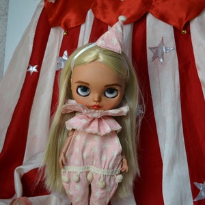 Op de afbeelding: Een blonde pop met een roze en wit clownskostuum met een witte hoed en een zilveren hoofdband. De pop staat voor een rood-wit gestreepte stoffen achtergrond.