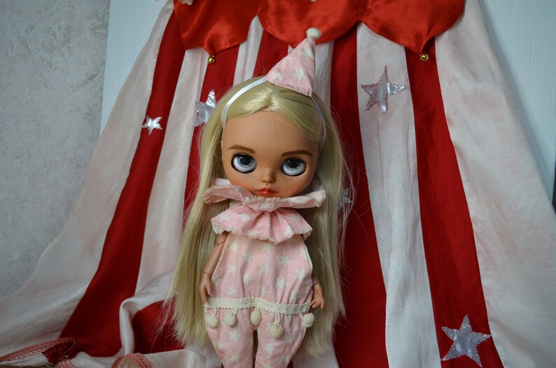Op de afbeelding: Een blonde pop met een roze en wit clownskostuum met een witte hoed en een zilveren hoofdband. De pop staat voor een rood-wit gestreepte stoffen achtergrond.