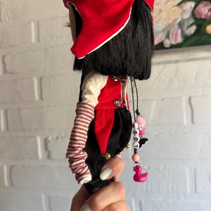 Pu&ograve; includere: Una bambola con capelli neri che indossa un cappello rosso, una maglia a maniche lunghe color crema con maniche a righe rosse e bianche, un gilet rosso con bottoni argentati e pantaloni neri. Un ciondolo di perline con la scritta LOVE e un'anatra rosa.