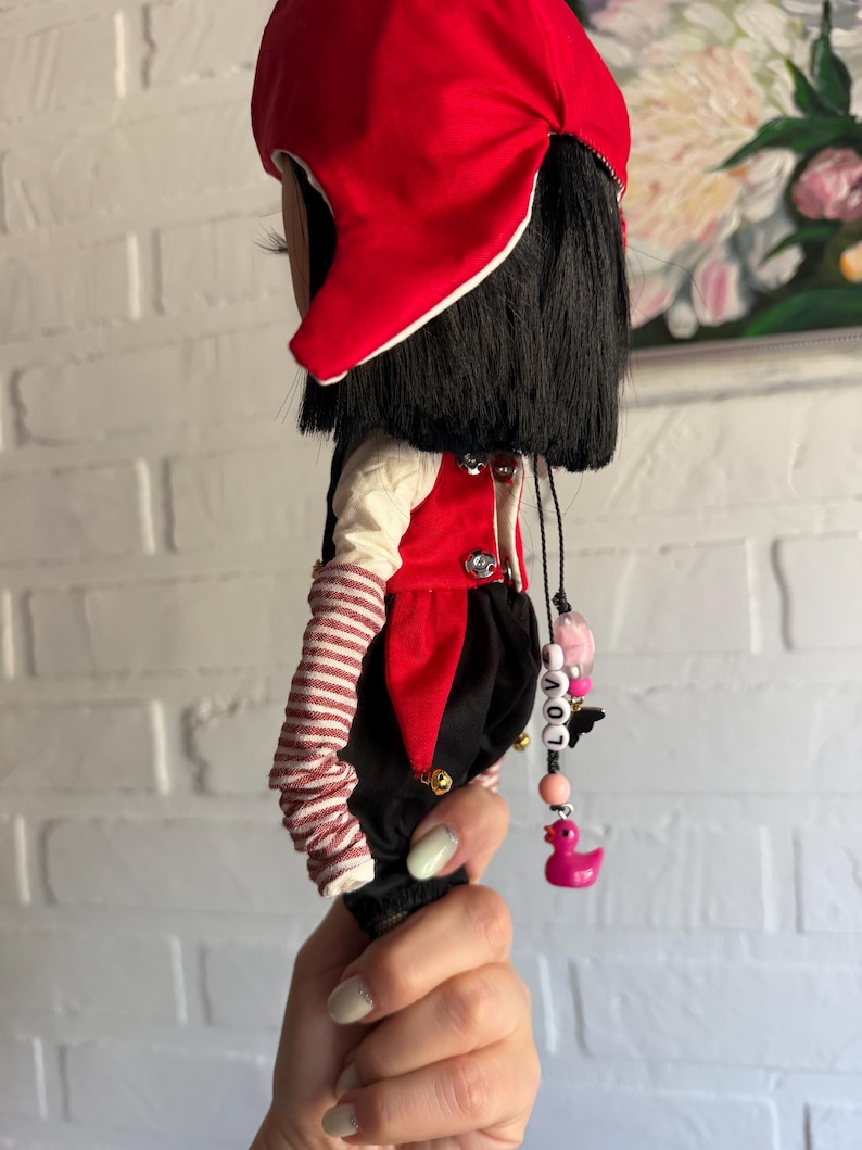Pu&ograve; includere: Una bambola con capelli neri che indossa un cappello rosso, una maglia a maniche lunghe color crema con maniche a righe rosse e bianche, un gilet rosso con bottoni argentati e pantaloni neri. Un ciondolo di perline con la scritta LOVE e un'anatra rosa.