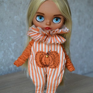 May include: A doll with blonde hair wearing an orange and white striped jumpsuit with a pumpkin design. The doll has blue eyes and is wearing brown shoes.