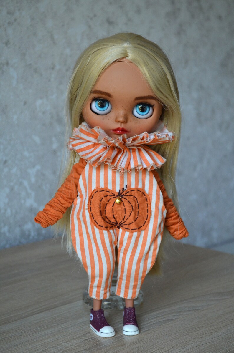 May include: A doll with blonde hair wearing an orange and white striped jumpsuit with a pumpkin design. The doll has blue eyes and is wearing brown shoes.
