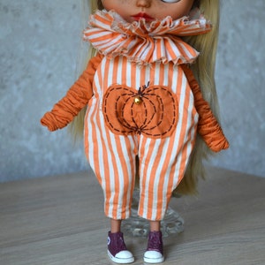 May include: A doll wearing an orange and white striped jumpsuit with a pumpkin design. The jumpsuit has a ruffled collar. The doll is wearing maroon shoes.