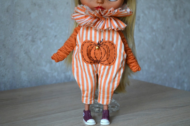 May include: A doll wearing an orange and white striped jumpsuit with a pumpkin design. The jumpsuit has a ruffled collar. The doll is wearing maroon shoes.