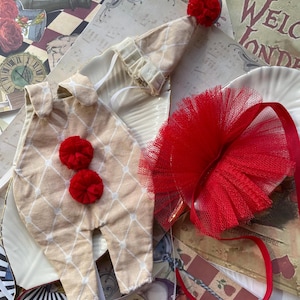Könnte beinhalten: Ein beige-weißer, gemusterter Overall mit roten Bommeln, ein roter Tüll-Tutu und ein beige-weißer, gemusterter Hut mit einer roten Bommel.