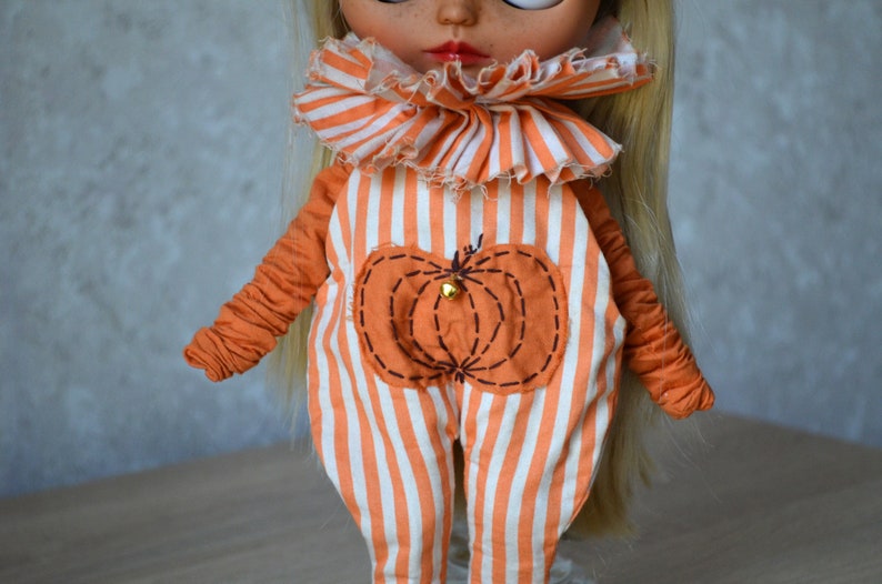 May include: A doll wearing an orange and white striped jumpsuit with a pumpkin design on the front. The jumpsuit has a ruffled collar.