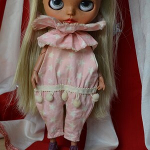 Op de afbeelding: Een roze en witte jumpsuit met een sterpatroon, een gerimpelde kraag en witte pompoenen aan de zoom. De pop draagt de jumpsuit en heeft blond haar.