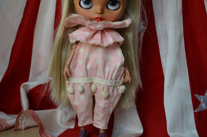 Op de afbeelding: Een roze en witte jumpsuit met een sterpatroon, een gerimpelde kraag en witte pompoenen aan de zoom. De pop draagt de jumpsuit en heeft blond haar.
