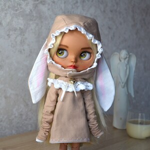 Puede incluir: Una mu&ntilde;eca con un vestido beige con capucha y orejas de conejo. El vestido tiene encaje blanco y una campana en la parte delantera. La mu&ntilde;eca tiene el pelo rubio y los ojos azules.