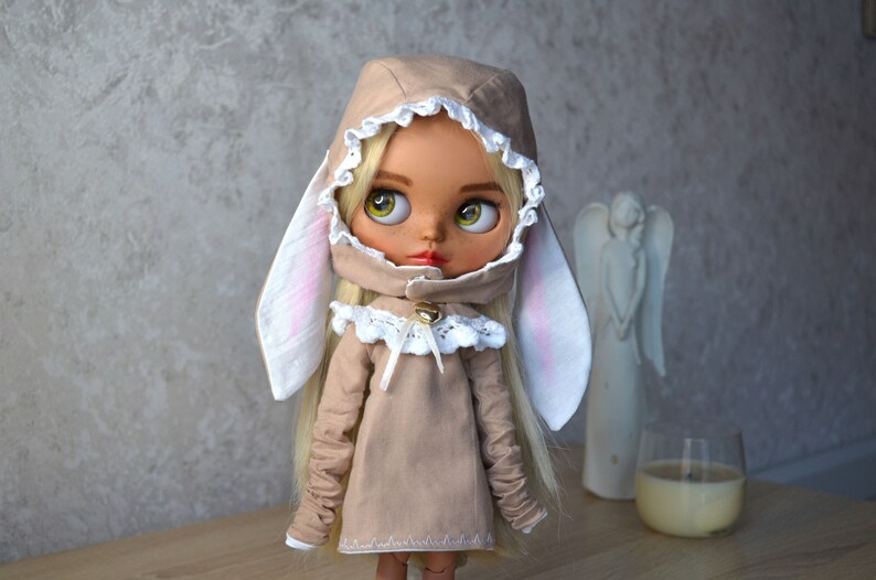 Puede incluir: Una mu&ntilde;eca con un vestido beige con capucha y orejas de conejo. El vestido tiene encaje blanco y una campana en la parte delantera. La mu&ntilde;eca tiene el pelo rubio y los ojos azules.