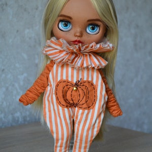May include: A doll with long blonde hair wearing an orange and white striped jumpsuit with a pumpkin patch on the front. The doll is wearing maroon trainers with white laces.