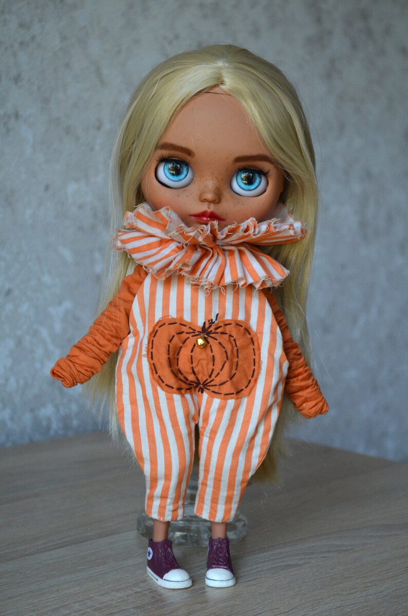 May include: A doll with long blonde hair wearing an orange and white striped jumpsuit with a pumpkin patch on the front. The doll is wearing maroon trainers with white laces.