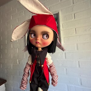 Pu&ograve; includere: Una bambola con capelli neri e occhi rosa indossa un cappello da giullare rosso e bianco con campanellini dorati. La bambola veste un gilet nero e rosso sopra una camicia bianca con maniche a righe, decorata con piccole stelle dorate e campanellini.