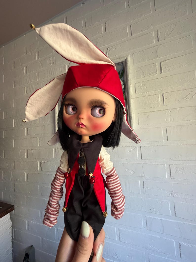 Pu&ograve; includere: Una bambola con capelli neri e occhi rosa indossa un cappello da giullare rosso e bianco con campanellini dorati. La bambola veste un gilet nero e rosso sopra una camicia bianca con maniche a righe, decorata con piccole stelle dorate e campanellini.