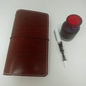 May include: A brown leather journal with white stitching and a rubber band closure. A white fountain pen and a red glass bottle of ink are also pictured.