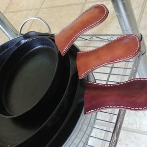May include: A stack of black cast iron skillets with three brown leather handle covers. The handle covers are stitched with white thread. The skillets are oval-shaped and nested together.