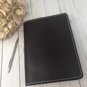 May include: A dark brown leather journal with white stitching around the edges. A silver pen with gold accents rests beside the journal. Dried flowers are in the background. The journal is on a light wood surface.