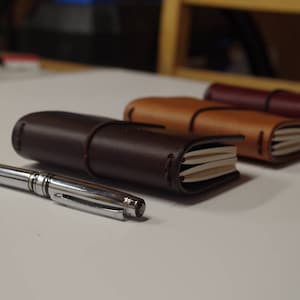May include: Three leather-bound journals in brown, tan, and burgundy, closed and stacked on a white surface. A silver pen is lying in front of the journals.