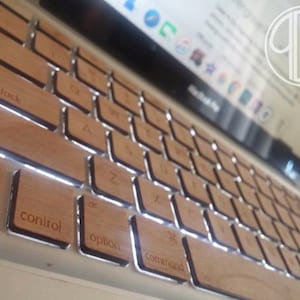 May include: A close-up of a laptop keyboard with wooden keycaps. The keys are labeled with the standard letters, numbers, and symbols. The keyboard is lit up, and the keys are reflecting the light.