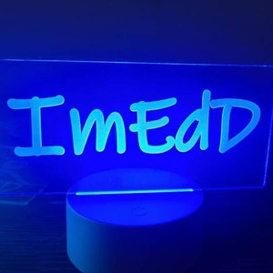 May include: A blue-lit LED sign with the word "ImEdD" in a handwritten style. The sign is rectangular and sits on a white, circular base. The background is dark, enhancing the glowing effect of the sign.