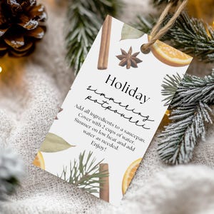 May include: A white tag with the words "Holiday simmering potpourri" in black script. The tag includes instructions and illustrations of cinnamon sticks, star anise, orange slices, and pine branches. The tag is tied with twine.