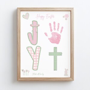 Könnte beinhalten: Ein gerahmter Druck mit einem hellen Holzrahmen. Der Druck zeigt die Worte "Happy Easter" in rosa Schrift, zusammen mit den Buchstaben "J" und "Y", einem Handabdruck und einem Kreuz. Die Buchstaben und das Kreuz sind grün und rosa Vichy. Ein Herz, Blumen und ein Hase sind ebenfalls enthalten.