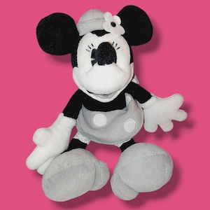 Op de afbeelding: Een Minnie Mouse pluche pop in zwart, wit en grijs. De pop heeft grote zwarte oren, een grijze hoed met een witte bloem en een grijze rok met witte stippen. De armen en voeten van de pop zijn wit.