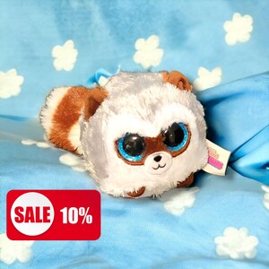 May include: A round plush toy resembling a raccoon with soft white and gray fur, large sparkling blue eyes, brown ear tips, and a striped tail. The plush features a small black nose and a stitched mouth, with a tag attached to the side.