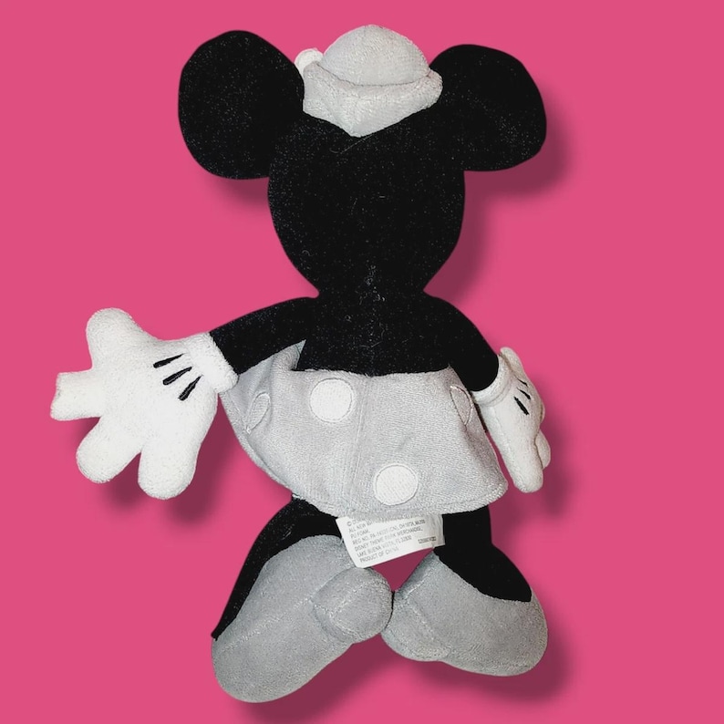 Op de afbeelding: Een pluche Minnie Mouse pop, van achteren gezien, met zwarte vacht, witte handschoenen en een grijze rok met witte stippen. De pop heeft een grijze hoed en grijze schoenen. De achtergrond is effen roze.