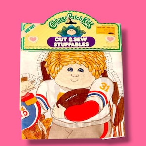 Może przedstawiać: Opakowanie vintage Cabbage Patch Kids Cut & Sew Stuffables. Na zdjęciu znajduje się ilustracja kreskówkowa Cabbage Patch Kid trzymającego piłkę nożną. Opakowanie jest białe, z logo Cabbage Patch Kids na górze i napisem "CUT & SEW STUFFABLES".