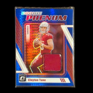 May include: A trading card featuring a football player in a red uniform, holding a football. The card has a blue and red design with the words "ROOKIE PHENOM" at the top. The player's name, Clayton Tune, is at the bottom.