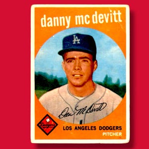May include: Vintage baseball card featuring Danny McDevitt of the Los Angeles Dodgers. The card has an orange background with a portrait of McDevitt wearing a Dodgers cap. The card includes his signature and team information.