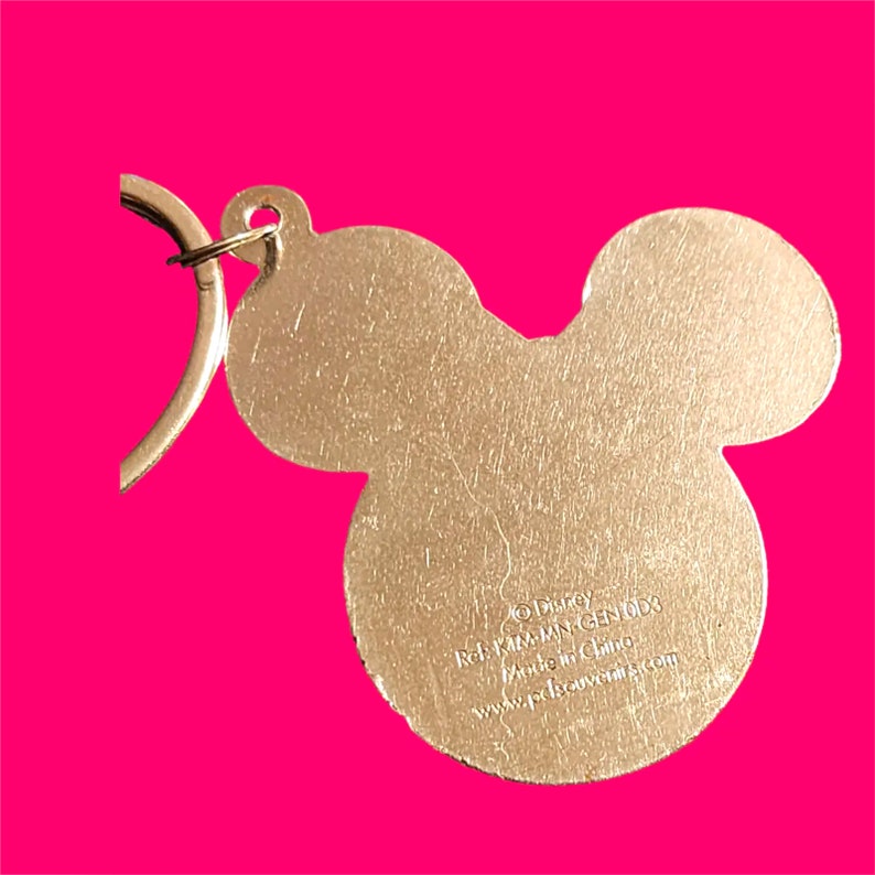 May include: A gold-colored Mickey Mouse keychain. The keychain is in the shape of Mickey Mouse's head and ears. The back of the keychain has text that reads "&copy; Disney, Ref. KIM-MAN, GEN 003, Made in China, www.pdssouvenirs.com."