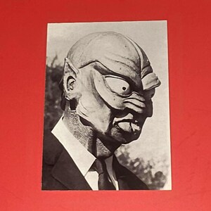 May include: Black and white photograph of a creature mask with large eyes, a prominent nose, and a textured surface. The mask is worn with a suit and tie, suggesting a formal setting. The image is set against a red background.