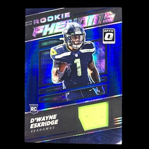 May include: A football trading card featuring D'Wayne Eskridge of the Seattle Seahawks. The card has a blue holographic background with the words "ROOKIE PHENOMS" at the top. The player is wearing a navy blue uniform with the number 1. The card also has the Optic logo.