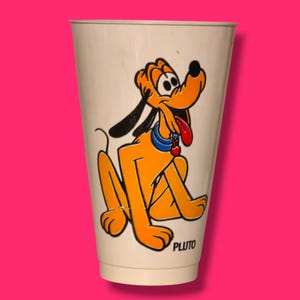 May include: A vintage off-white plastic cup featuring a cartoon illustration of Pluto, the Disney dog. Pluto is depicted in orange with black ears and a red tongue, wearing a blue collar. The word "PLUTO" is printed at the bottom.
